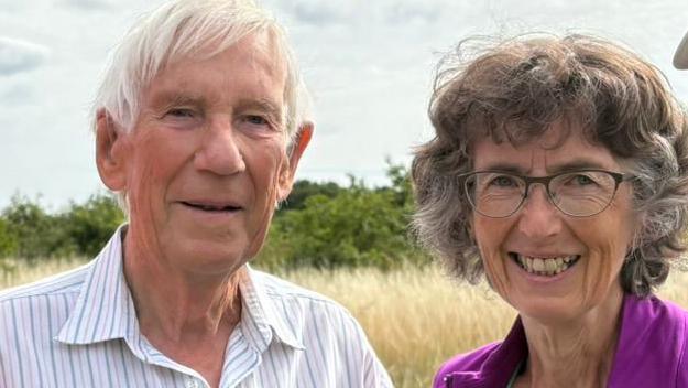 Michael Thorley has white hair and is wearing a light blue and white striped shirt. Next to him is his wife Sarah, wearing glasses and a purple top. They are standing in a field, smiling.