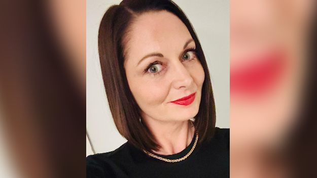Nicola wearing a dark top, with red lipstick and dark, short hair, smiling at the
camera.