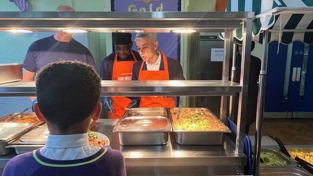 Sir Sadiq Khan is standing behind a glass hot food counter in a school as though he is serving food. He is standing alongside two staff members. He is looking down and appears to be talking.

A boy has his back the camera and faces the food counter as though waiting to be served.