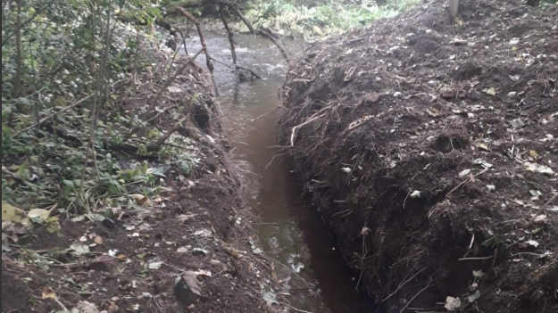 The image shows a channel dug out through a section of land with water running though it. Soil sits on either side and the main river can be seen in the background.