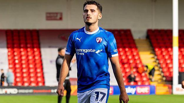 Chesterfield striker Armando Dobra in blue shirt and white shorts wandering on off the pitch after a warm-up with empty red seats behind him at Walsall