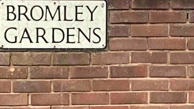 A white sign, with the words "BROMLEY GARDENS" written in black on a re-brick wall