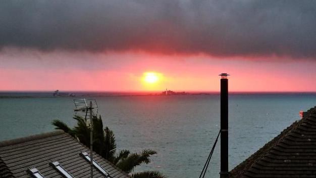 An orange sun at the centre of the image on the horizon, above the sea. It is sending out a reddish hew under very dark, heavy clouds above. In the foreground, in front of the sea are the roofs of two properties and a chimney.