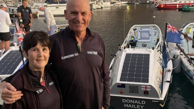 The couple are standing on a dock in purple tops next to their boat. Mr Parr has his arm around his wife.