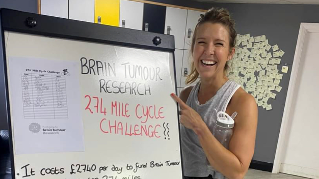 Ms Wrathall smiles at the camera, wearing a grey vest and carrying a water bottle. She is standing beside a movable white board that reads: Brain Tumour Research, 274 Mile Cycle Challenge. 