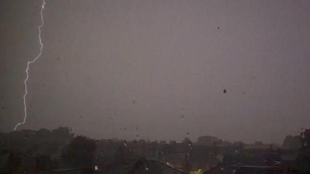 A lightning bolt strikes down over a row of houses during heavy rain, with dark skies and low visibility across a residential area.