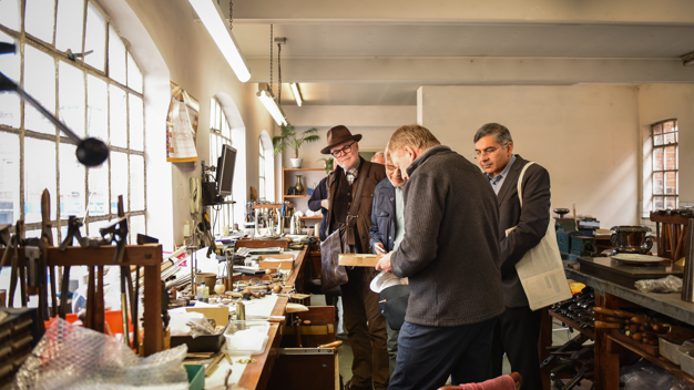 A group of men stand in a jewellery-making studio, looking down at an object in one of the men's hands. They stand next to a work bench with lots of tools on it.