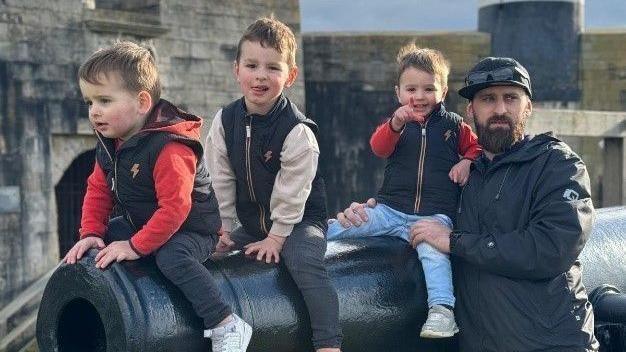 Three young children sit on a cannon, with a bearded man in a cap holding onto the youngest. 