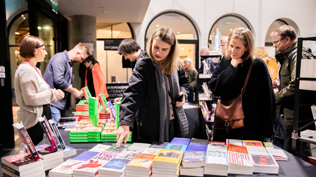 A queue of people look at piles of books on a table with a black tablecloth on it.