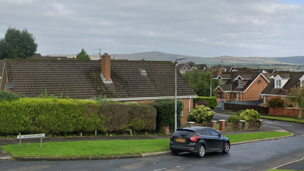 A residential area with brown houses with black roofs. There is a footpath with grass on one side and a hedge on the other. A black car is parked along the road as well. There is a white road sign with the name of the park written on it.