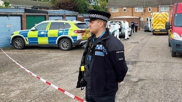 A police officer standing behind a cordon in a residential area. There is a police car and ambulance parked behind him, with forensic officers probing the scene.