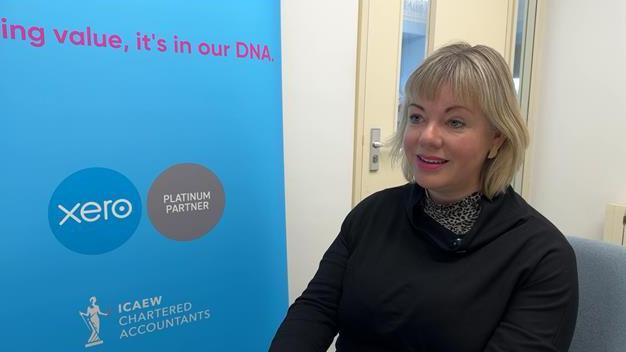 Director Diane de Garis is wearing a black top and is sat on a chair. She has short blonde hair and is sat next to a blue business sign. 