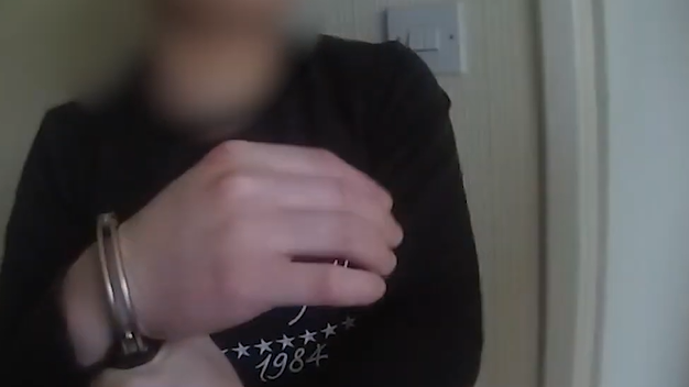 A person wearing a dark long-sleeve shirt is shown indoors with their hands restrained by metal handcuffs. The individual’s face is blurred for anonymity. The background includes a light-coloured wall, a doorframe, and a visible light switch.