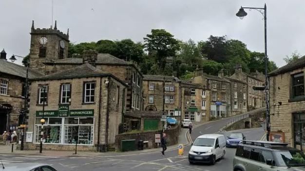 Cars drive through a crossroads in Holmfirth town centre which is edged by houses and shops in sooted West Yorkshire stone.