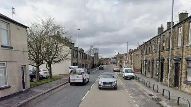 Rows of terraced houses line Rochdale Road on a cloudy day. There are two cars and a white van on the road in this Google Maps image. 