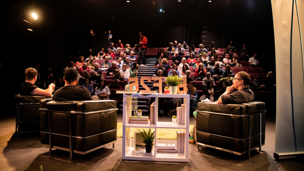 Three people in leather armchairs sit with their backs to the camera in front of an auditorium of people.
