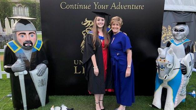 Grace in a cap and gown stands smiling with her mother, who wears a blue dress. They are standing outside in front of a black backdrop, with the wording printed "continue your adventure".