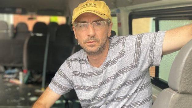 A man wearing a yellow cap and a grey t-shirt sitting in the back of a van.