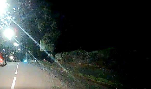 A roadway at night from the perspective of the driver looking onto a head lighted road with a stone wall along one side