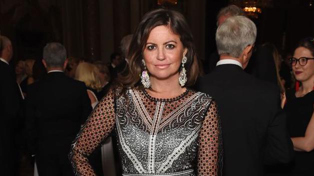 Dame Deborah James looks at the camera at a function where lots of smartly dressed people can be seen in the background. She is wearing an embroidered top with sheer sleeves and also large jewelled earrings and has long dark hair.