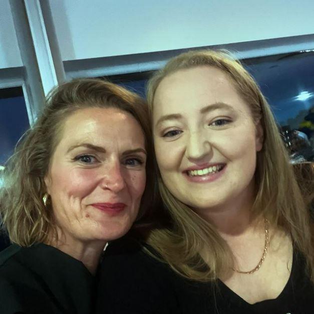 Two women standing close together in a room. The woman on the left has short blonde hair, a nosering and small gold hoop earrings. The woman on the right has long blonde hair and a gold necklace. They are both wearing black tops. There is a window in the background.