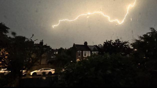 Lightning stretches across the sky above houses and a parked car during a heavy night storm.