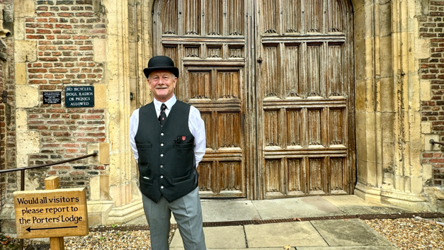 Dean Allen stood to the left of a large wooden double door which is set against a stone arch and red bricked walls. To the bottom left of the image is a small wooden sign which reads 'Would all visitors please reports to the Porter's Lodge'. Next to the sign is Mr Allen who has his hands behind his back and is standing to face the camera while smiling. He wears a porter's outfit which consists of a black bowler hat, white shirt, black tie, black waistcoat and grey suit trousers. The black tie and breast pocket of the waist coat have the Jesus College logo. This is a shield shape with a red colouring and golden crowns around the edges. The centre is white with a black horizontal line in the middle. Above this line are two images of cockerel heads, there is a third one below the black line. 