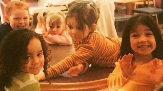 Five young girls look at the camera while sitting around a table at McDonald's in 1997. The picture has an orange tint. The girl on the right is waving at the camera and beaming, while the girl next to her is Alice Connor and is leaning across the table. They are made up to look like young versions of the Spice Girls. 