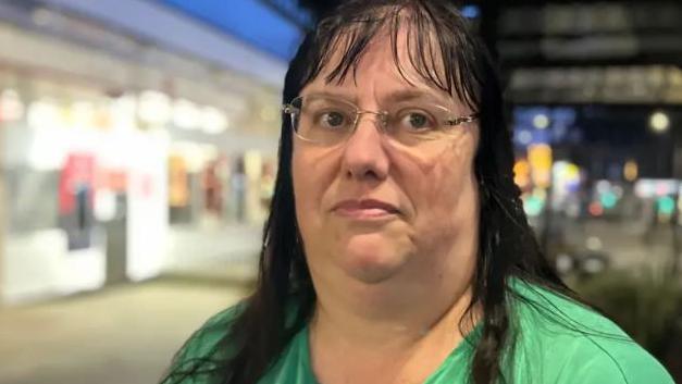 Ms Dominic wears glasses and a green blouse. She has long black hair and a fringe. The post office can be made out in the background.