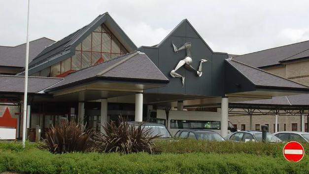 The front entrance to Noble's Hospital. A row of cars is parked in front of a canopy featuring a large silver three-legs-of-Mann emblem, which is held up by white pillars. The roofs of the building are sloped and there are a no entry sign and a white flagpole visible above a hedgerow in front of the cars.