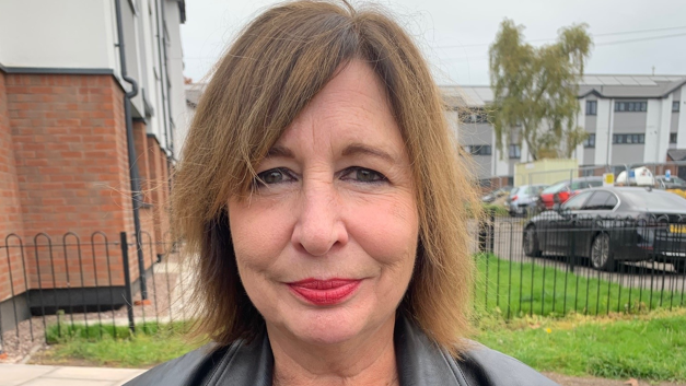 Ms Francis is stood outside the flats facing the camera and smiling. She has make up on and short, brown hair and is wearing a black jacket. 