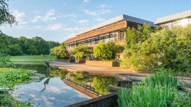View taken of the HQ building with a pond in front of the building. There are vegetation and trees surrounding the site. The building itself is made up from a mix of concrete and glass and is at least two storeys.