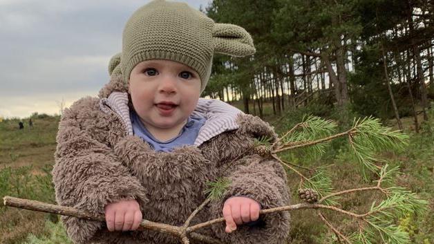 A baby dressed in woollen clothes and a pink woollen hat is holding a pine tree branch in a nature reserve. Other people could be seen in the background. It is overcast.