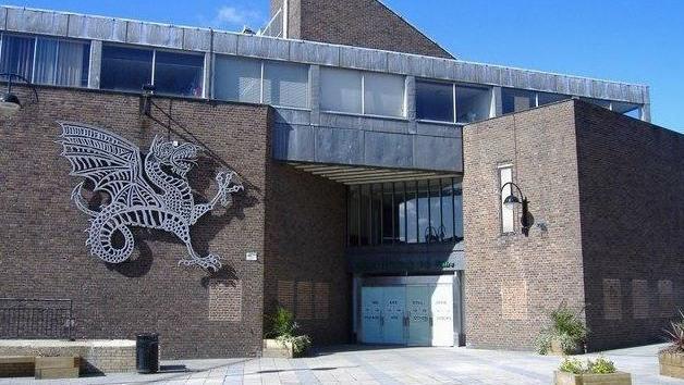 The exterior of Wyvern Theatre, pictured on a sunny day. The building is built of dark brick and appears dated, with gaps where poster boards may have been and weathering to the roos of the building. A large metal sculpture of a dragon is attached to the outside of the building, to the left of the doors. 