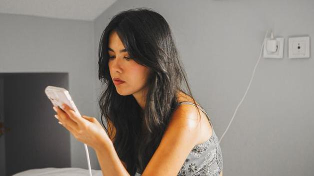A woman uses a smartphone while charging it in a bedroom. She is sitting on a bed and the phone cable is plugged into a wall socket behind her. She has long black hair and is wearing a top with straps, and she is sitting looking at the screen.