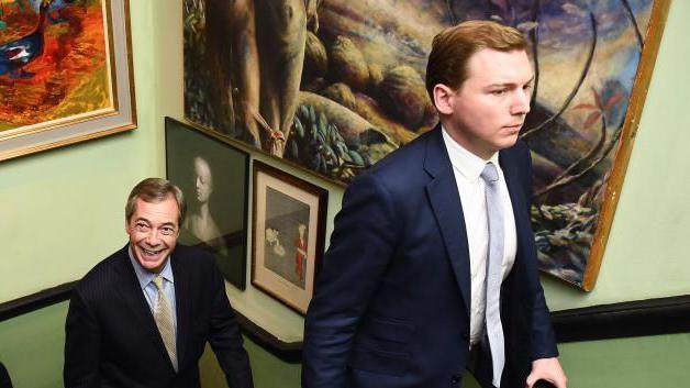 George Cottrell, in suit and tie, walks up a staircase past a wall decorated with pictures with Nigel Farage, also in suit and tie, following