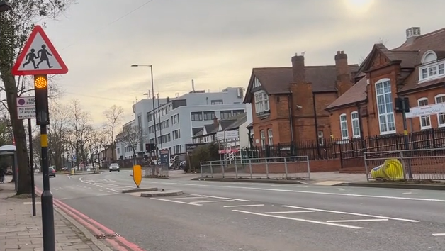 A road with a red and white children crossing warning sign in the foreground. There is a large red brick building on the opposite side of the road as well as a large white building. 