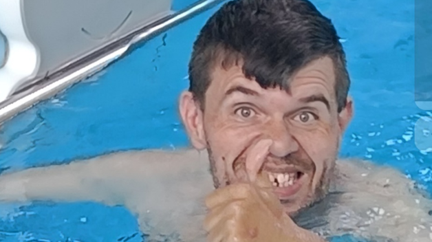 A white man with brown hair and stubble does a thumbs up gesture while swimming in a swimming pool
