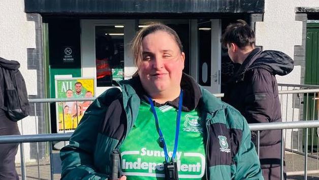 A picture of Jo stood in front of the Plymouth Argyle superstore. She is wearing a green Argyle top and green Argyle coat.