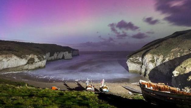 View of boats on the beach at Flamborough, flanked by cliffs, with the green and purple of the Northern Lights in the background