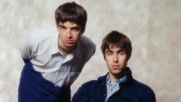Noel, crouching down, next to Liam sitting on a chair, unsmiling looking directly at the camera.
