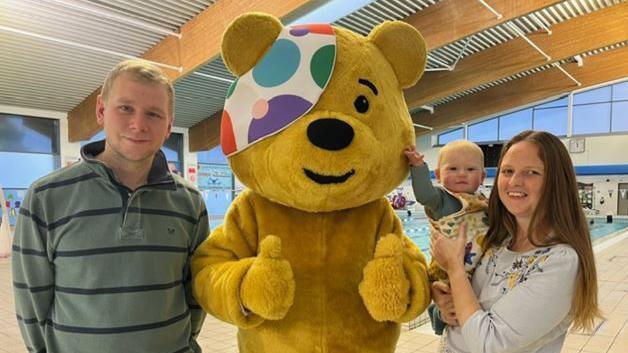 Parents Simon and Tammy smiling and standing either side of Pudsey, who is putting both thumbs up. Pudsey is a yellow bear wearing a colourful eye patch. Tammy is holding baby Everett who is looking at the camera and touching Pudsey's cheek. Simon is wearing a green and black striped jumper and Tammy has long brown hair and is wearing a white top with flowers on. They are standing in front of a pool.