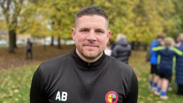 A man wearing a black training jacket stood in front of a green space and people in the background