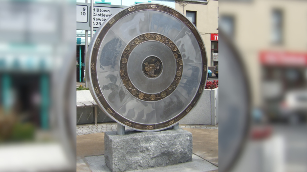 A large grey, bronze and circular piece of artwork with an old Irish penny in the middle. It is positioned on a stone block and behind it is flower beds and shopfronts with sign posts. There are various symbols etched onto the artwork to represent various aspects of the market.