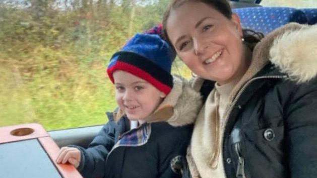 A woman sitting on a train with her young son. The woman has brown hair and is smiling. She has a cream coloured top and a black coat. The boy is wearing a navy coat and a blue, black and red hat. 