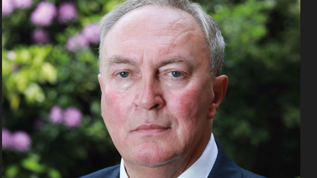 A white man with grey hair stares at the camera. He is wearing a blue suit jacket, white shirt and red tie.