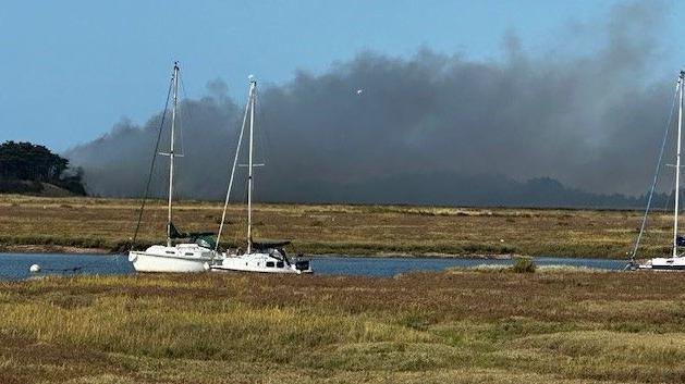 A cloud of black smoke rising in the sky above scrubby land three white yachts moored on water in the foreground