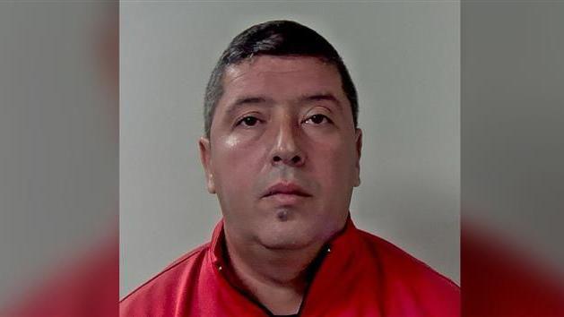 A police mugshot of Marius Bajenaru. He has black hair and is wearing a red jacket.