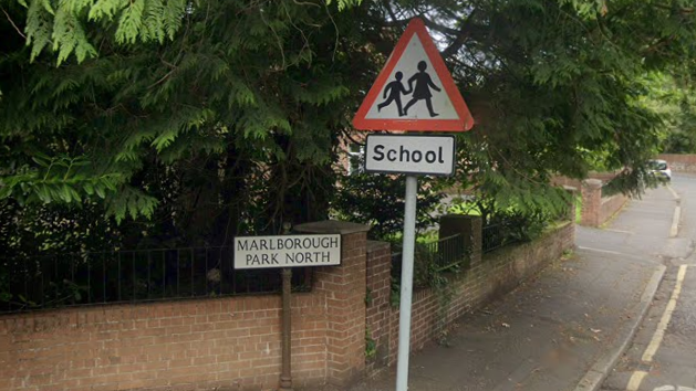 A Google Maps screenshot of Marlborough Park North. A small road sign displaying the road name is place on a short pole against a small red brick wall and green trees. To the right of the road sign is a triangle school sign showing school children.