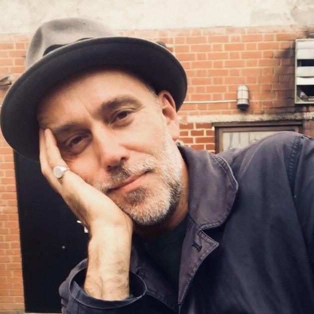 A man smiles at the camera, with his head to one side, learning on his hand, he has a hat on and is standing in front of a brick wall.